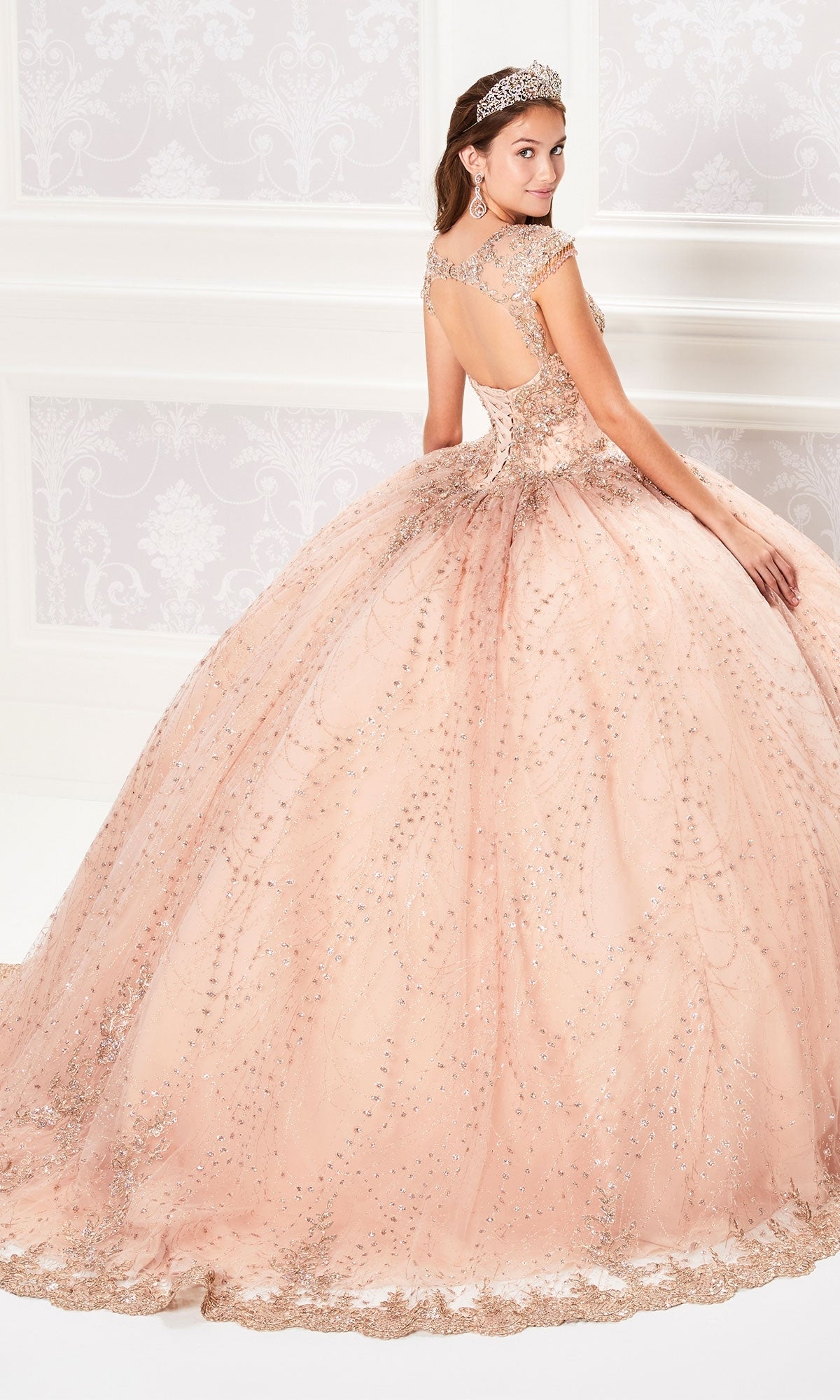 A woman in the Princesa PR21964 Cap-Sleeve Quinceanera Dress—sparkling, peach-colored with a full skirt and lace applique—stands elegantly in a room, facing away and glancing over her shoulder.